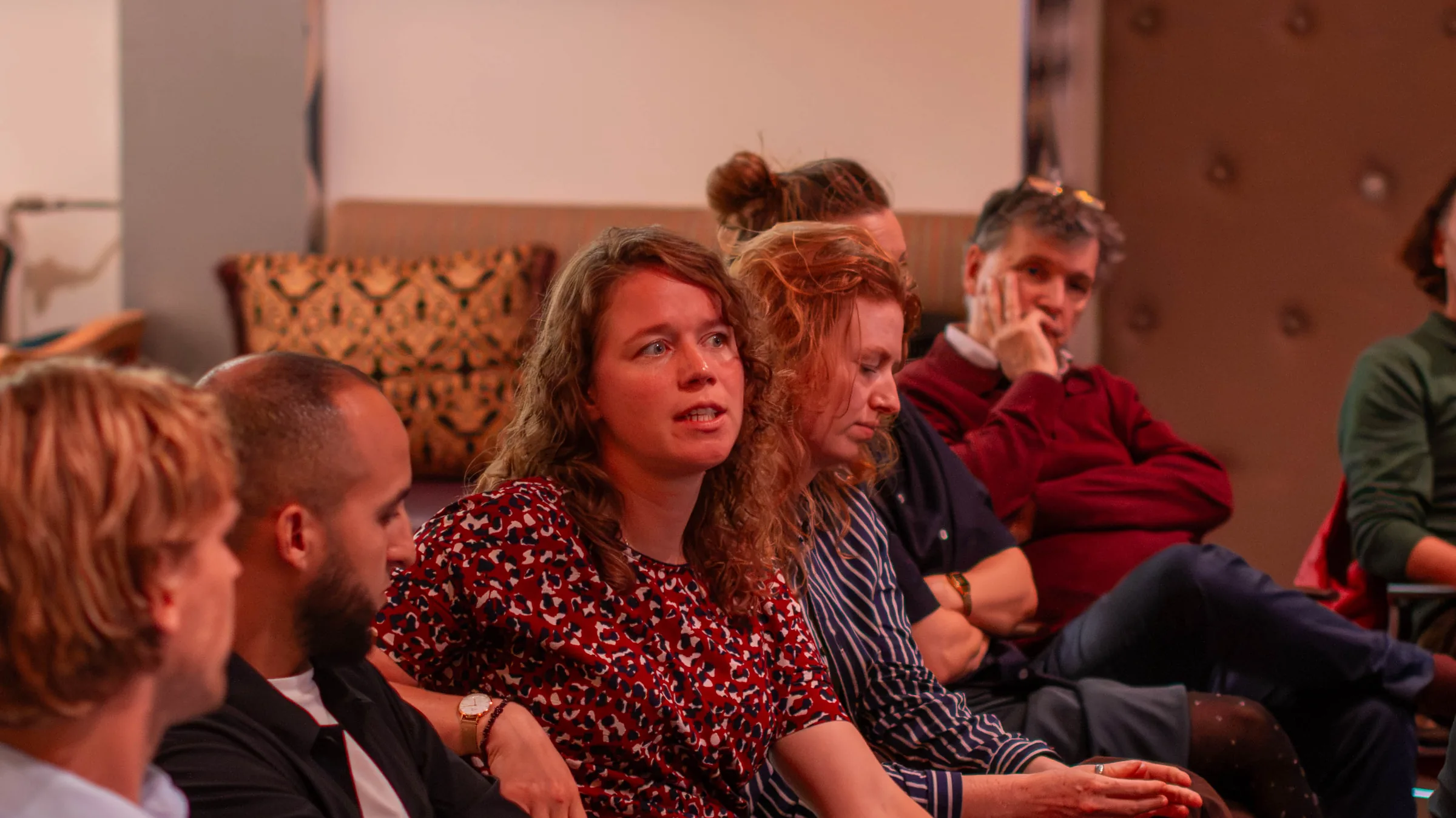Een deelnemer met krullend haar neemt het woord tijdens de bijeenkomst; om haar heen luisteren andere deelnemers.