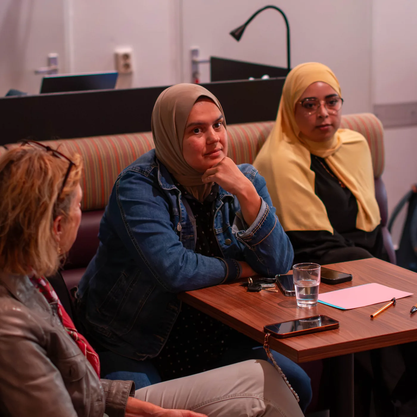 Twee jonge vrouwen met hoofddoek aan tafel in gesprek met een andere deelnemer.